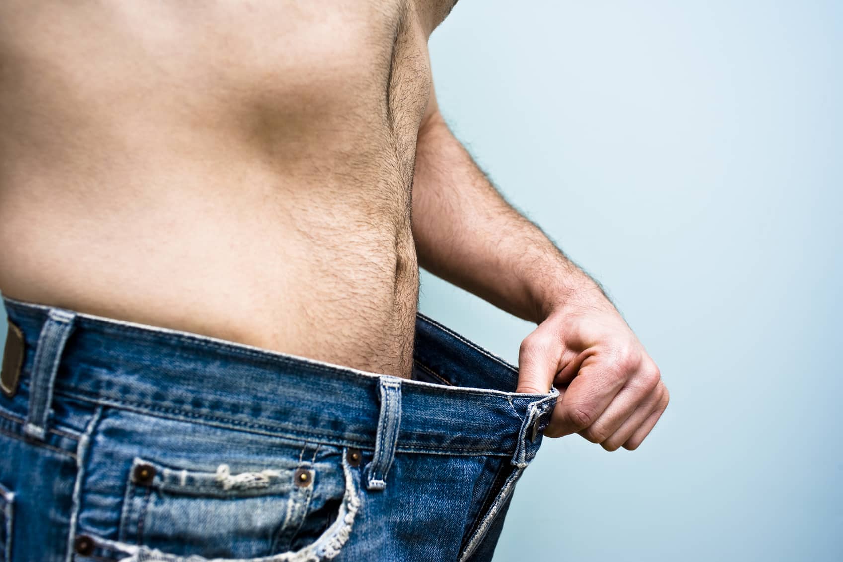 Man stands with an old pair of blue jeans showing his recent weight loss