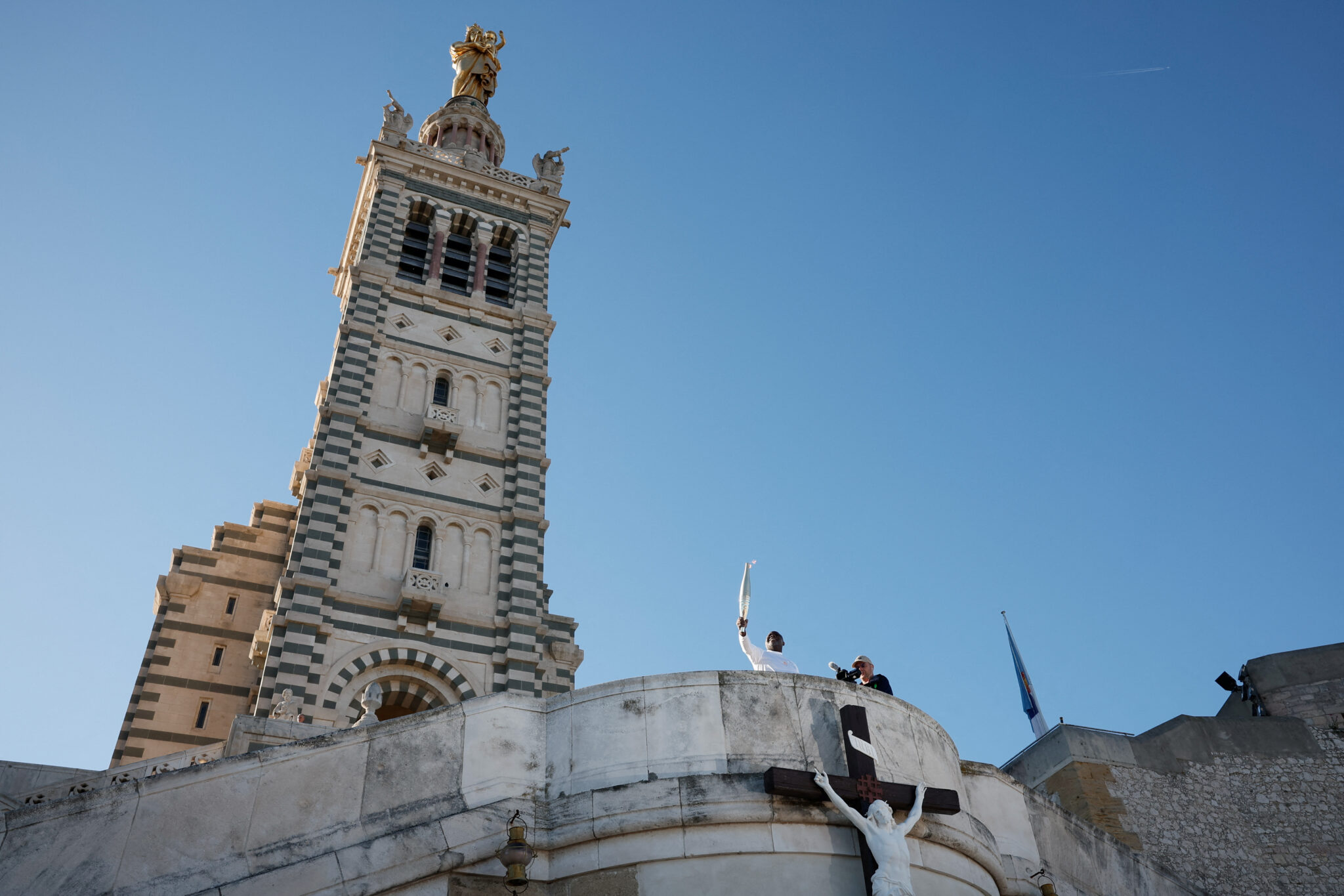 Torch bearer French former football player Basile Boli holds the Olympics torch with Notre-Dame de la Garde Basilica in the background during the relay ahead Paris 2024 Olympic games in Marseille, France, May 9, 2024. REUTERS/Benoit Tessier