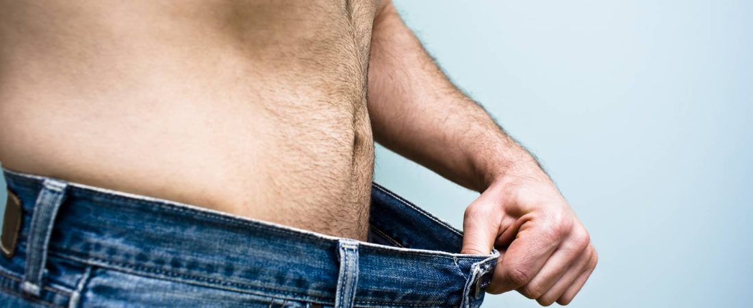 Man stands with an old pair of blue jeans showing his recent weight loss