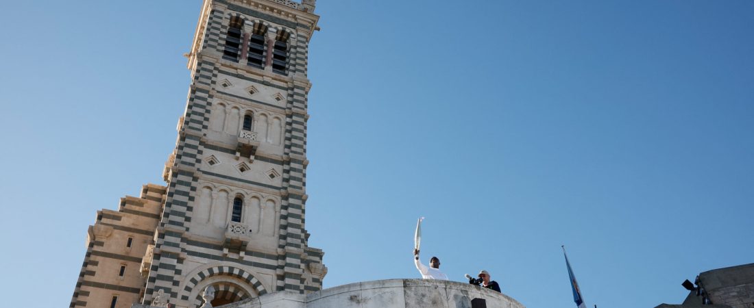 Torch bearer French former football player Basile Boli holds the Olympics torch with Notre-Dame de la Garde Basilica in the background during the relay ahead Paris 2024 Olympic games in Marseille, France, May 9, 2024. REUTERS/Benoit Tessier